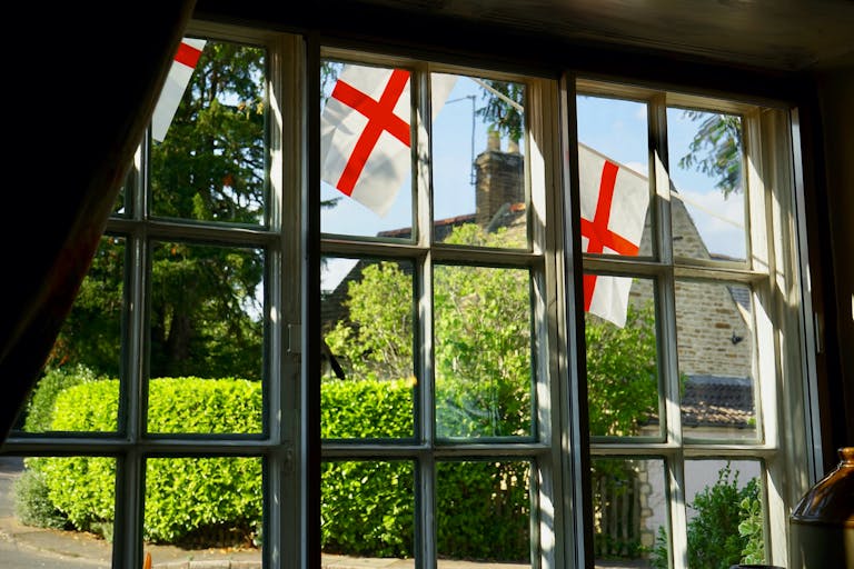 Quaint English cottage view through window with England flags outside, lush greenery visible.